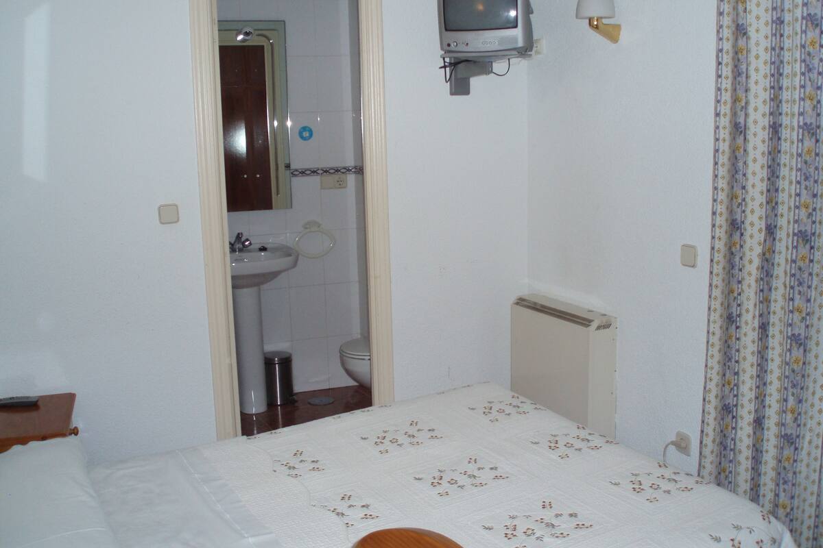 Ein kleines, schlichtes Zimmer mit Bett, Nachttisch, Wand-Fernseher, kleinem Heizkörper, Spüle und Toilette.