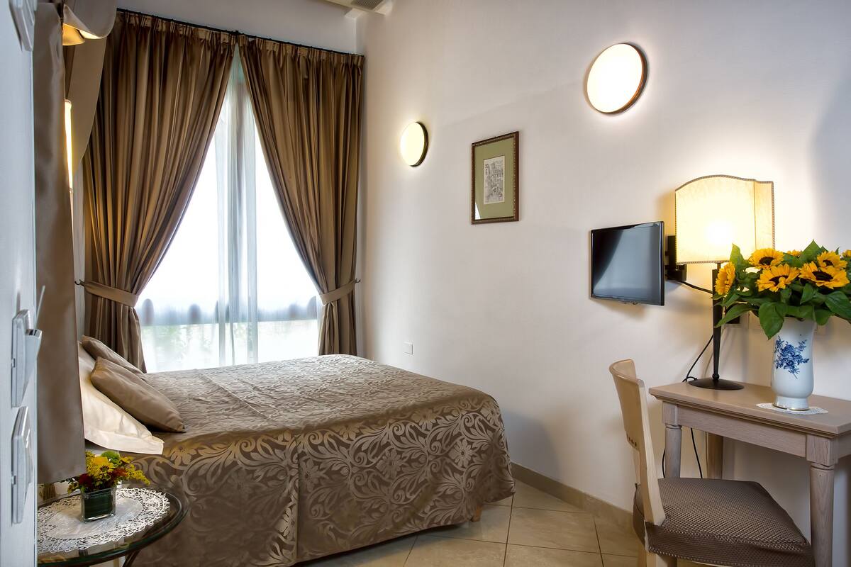 A hotel room with a bed, a desk with a lamp, a vase with flowers, and a television on the wall.