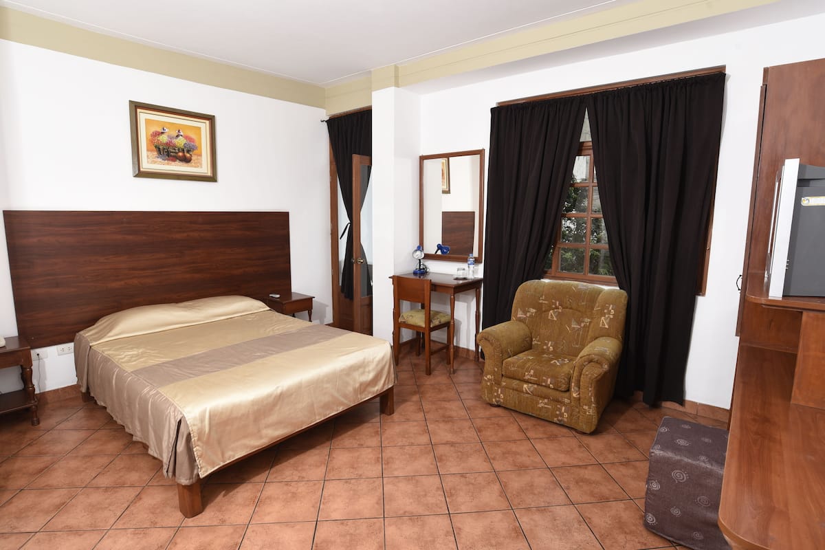 Quarto de hotel com cama, escrivaninha, cadeira, espelho e janela com cortinas.
