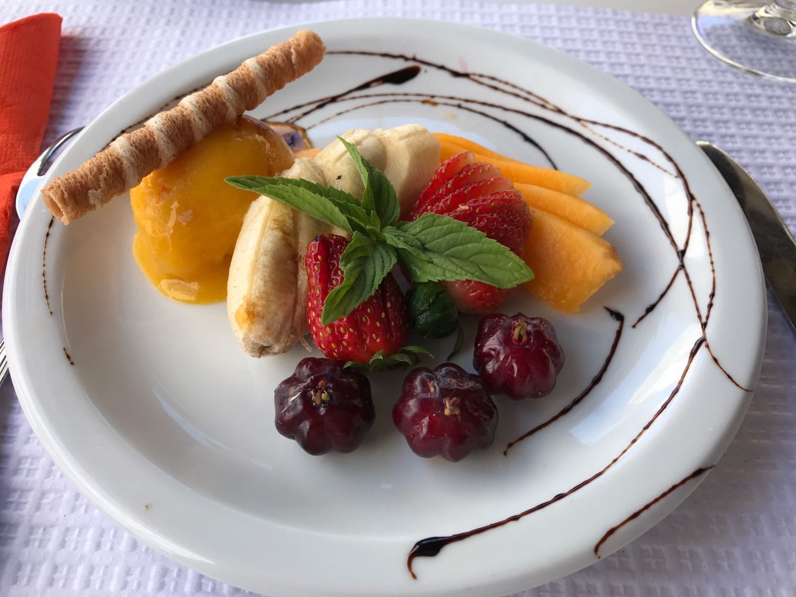 A plate with a banana, strawberries, grapes, and a slice of mango, garnished with mint leaves and chocolate drizzle.