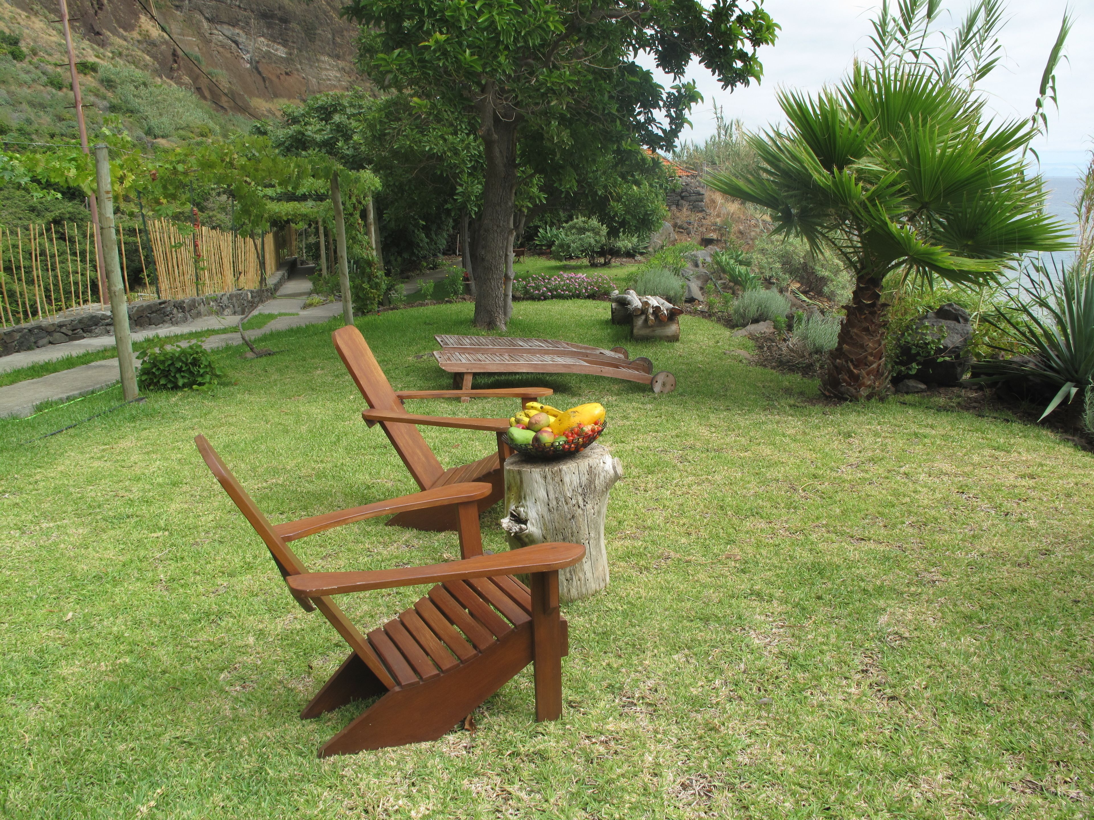 A grassy outdoor area with wooden chairs and a table with fruit.