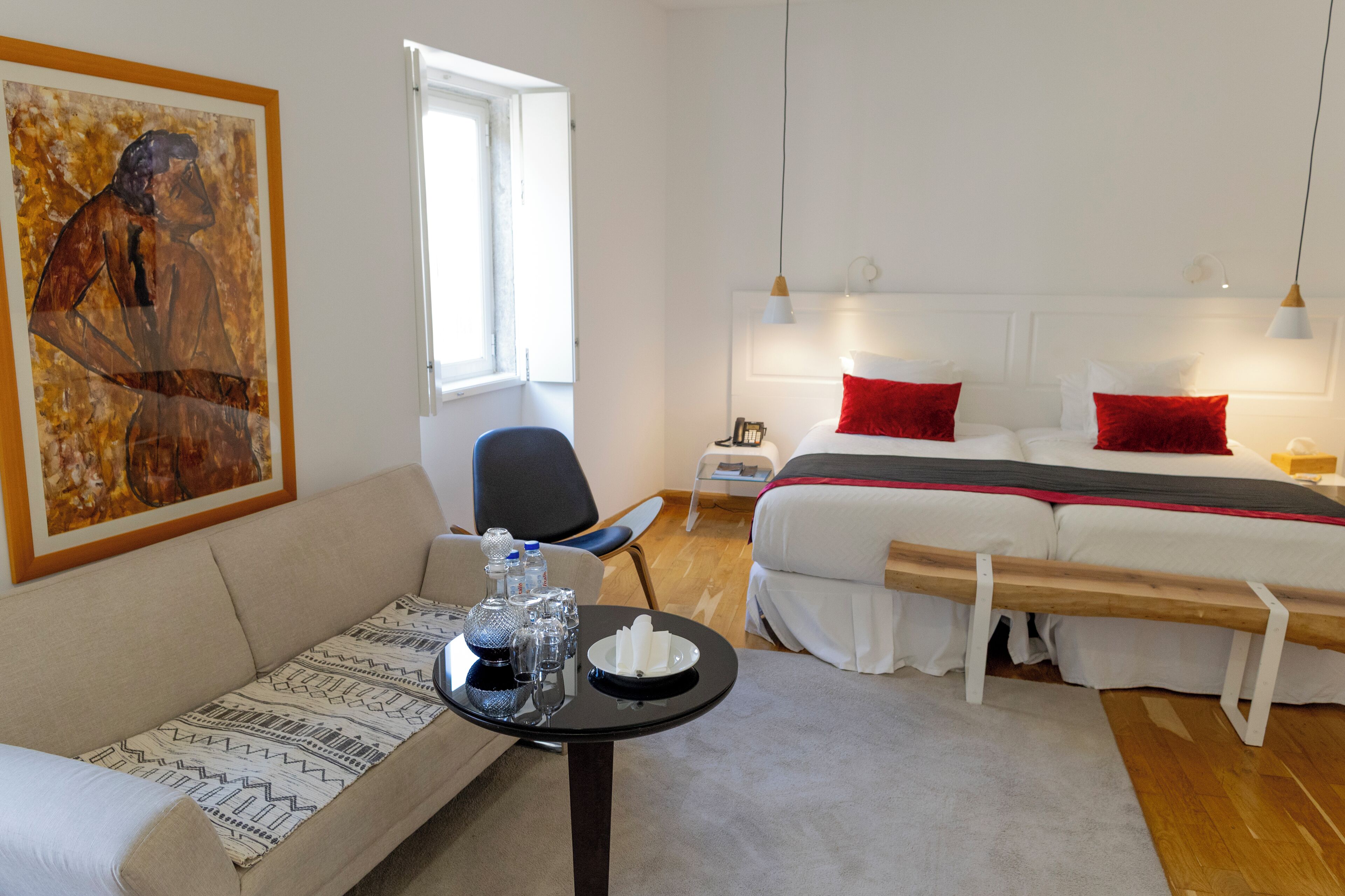 A hotel room with a sofa, a small table with a tray of glasses and a bottle, a bed with red pillows, and a painting on the wall.