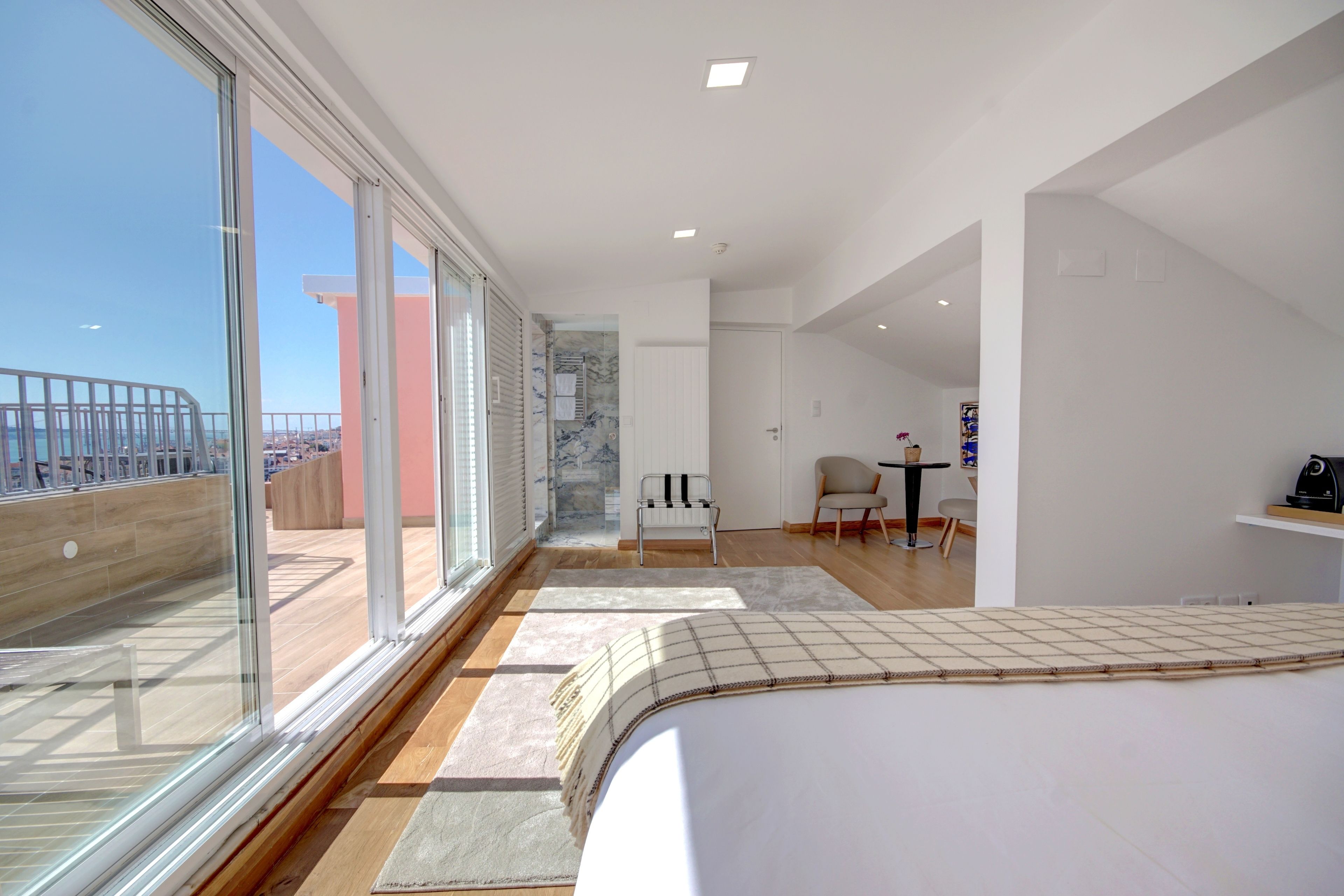 Ein modernes Hotelzimmer mit einem großen Balkon, einem Bett mit kariertem Überwurf und einer kleinen Sitzecke mit Stuhl und Tisch.