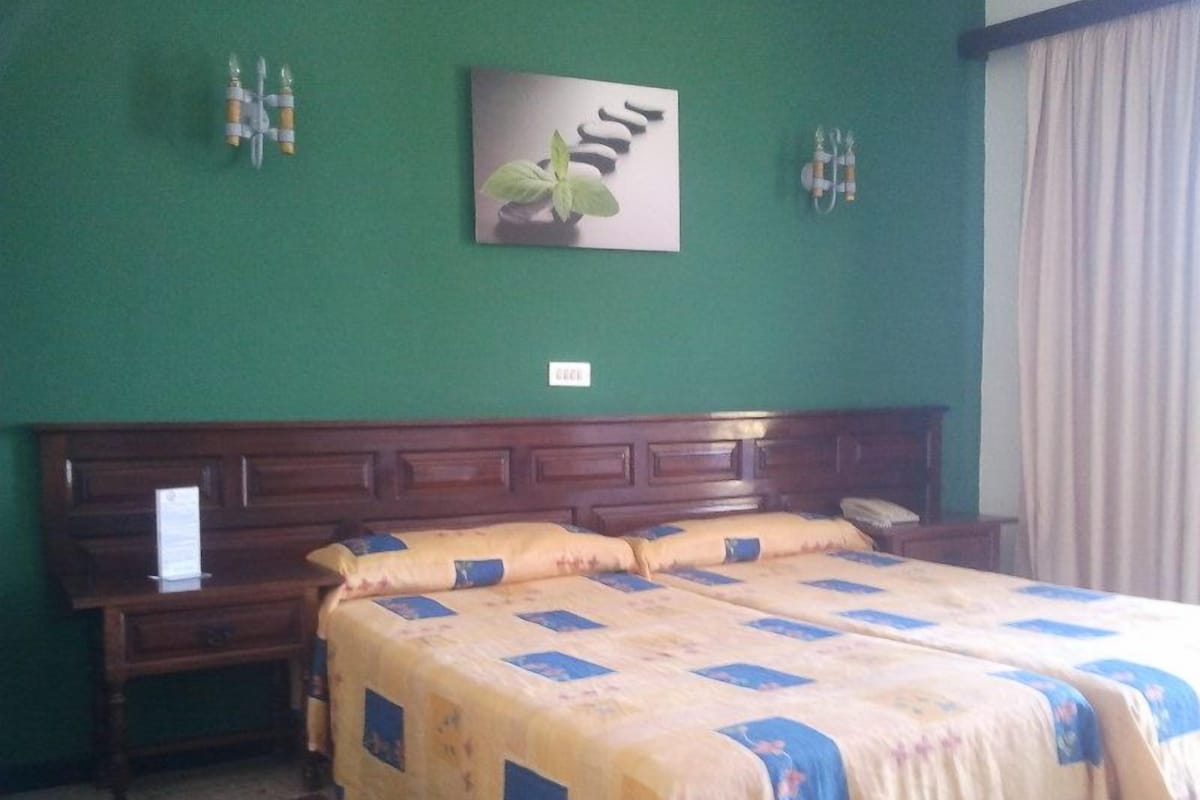 Quarto de hotel com uma cama grande, cabeceira de madeira, parede verde e uma janela com cortinas.