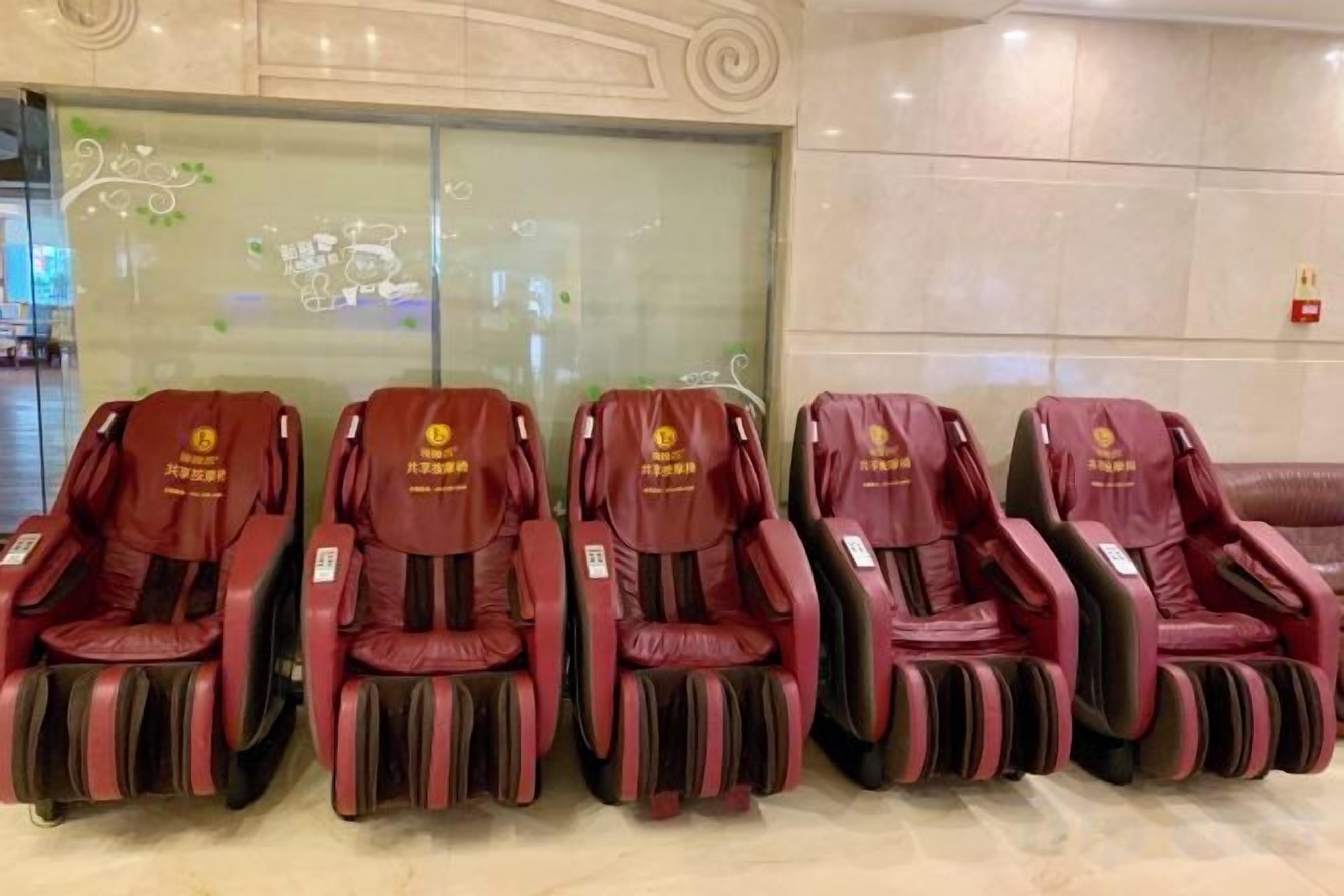 A row of red massage chairs in a lobby area with a glass wall and a tiled floor.