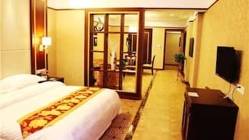 A hotel room with a large bed, a desk with a television, a mirror, and a sliding door leading to a dining area.