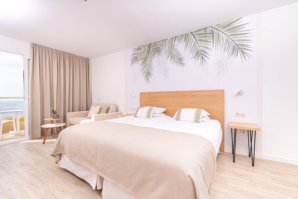 Une chambre d’hôtel moderne dotée d’un grand lit, d’un coin salon avec un fauteuil et une petite table, et offrant une vue sur l’extérieur.