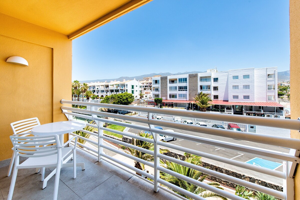 A balcony with white chairs and a table, overlooking a parking area and buildings with a clear sky.