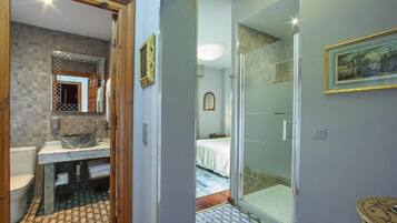 Un baño moderno con suelo a cuadros, cabina de ducha de vidrio y una cama visible en la habitación contigua.