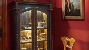 A room with a dark wooden cabinet displaying various items, a red wall with a framed painting, and a wooden chair.