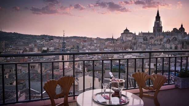 Un balcón con una mesa dispuesta para dos personas, con vistas a un paisaje urbano que incluye edificios históricos y una destacada torre de iglesia.