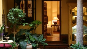 Un interior tradicional con una puerta de madera que da a una habitación con una cama y una lámpara. Hay plantas en macetas y una mesita con un florero.