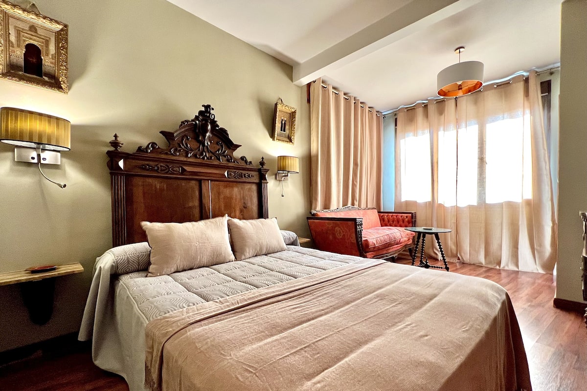 A hotel room with a large bed, a wooden headboard, a red armchair, a small table, and a large window with curtains.