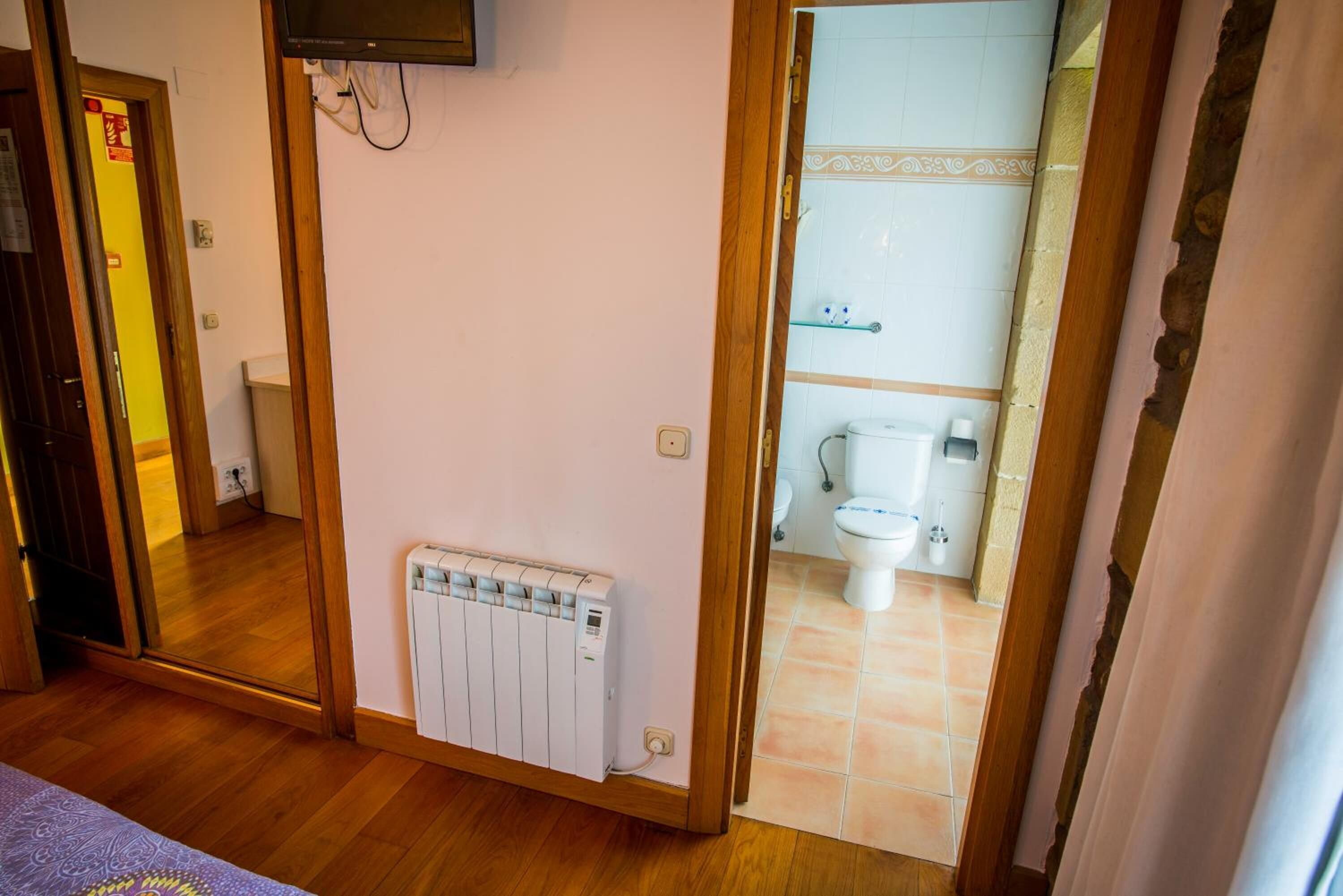A room with a wooden floor, a radiator, a television mounted on the wall, and a bathroom visible through an open door.