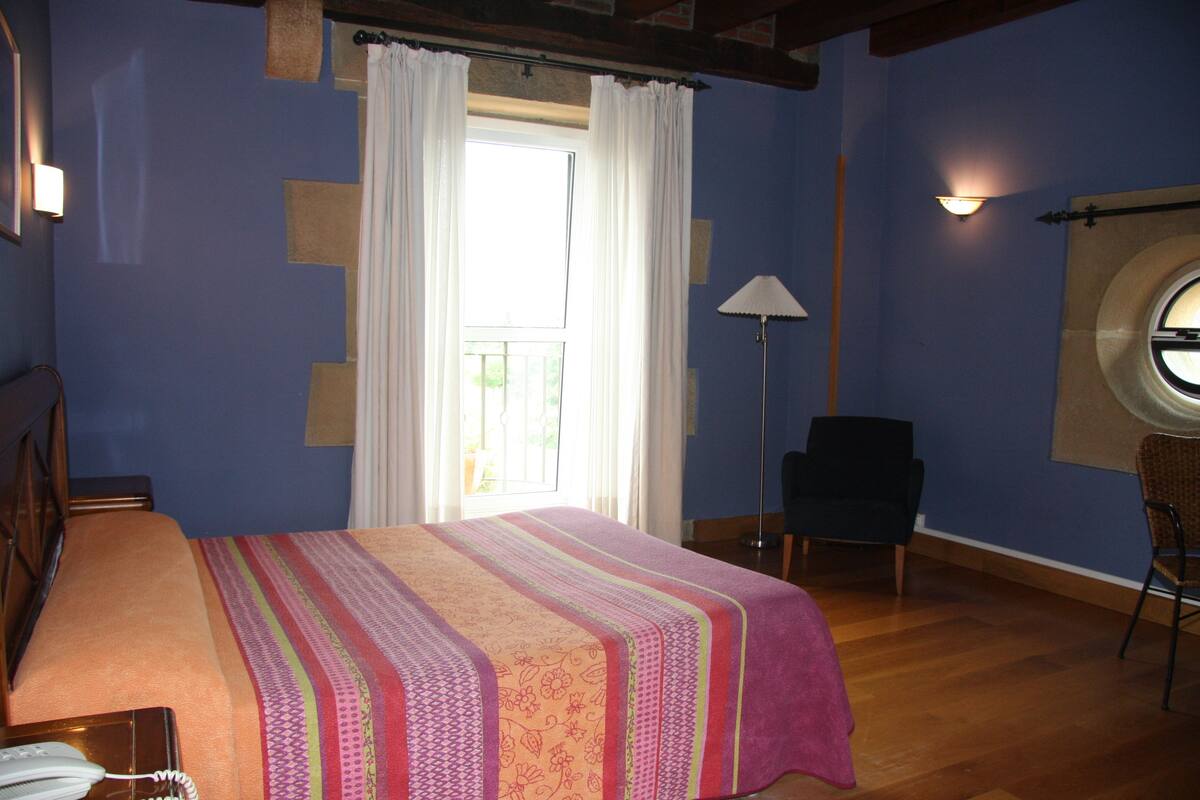 A bedroom with a bed, a chair, a lamp, a window, and a wall-mounted light fixture.