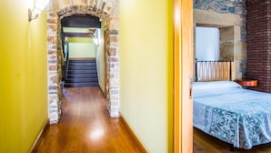 A narrow hallway with a stone archway leading to a staircase, a bed with a patterned bedspread, and a stone wall with a window. - Hotel Palacio Atxega (Usurbil)