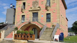 A pink building with a central archway, a staircase, and a balcony with a metal railing. - Hotel Palacio Atxega (Usurbil)