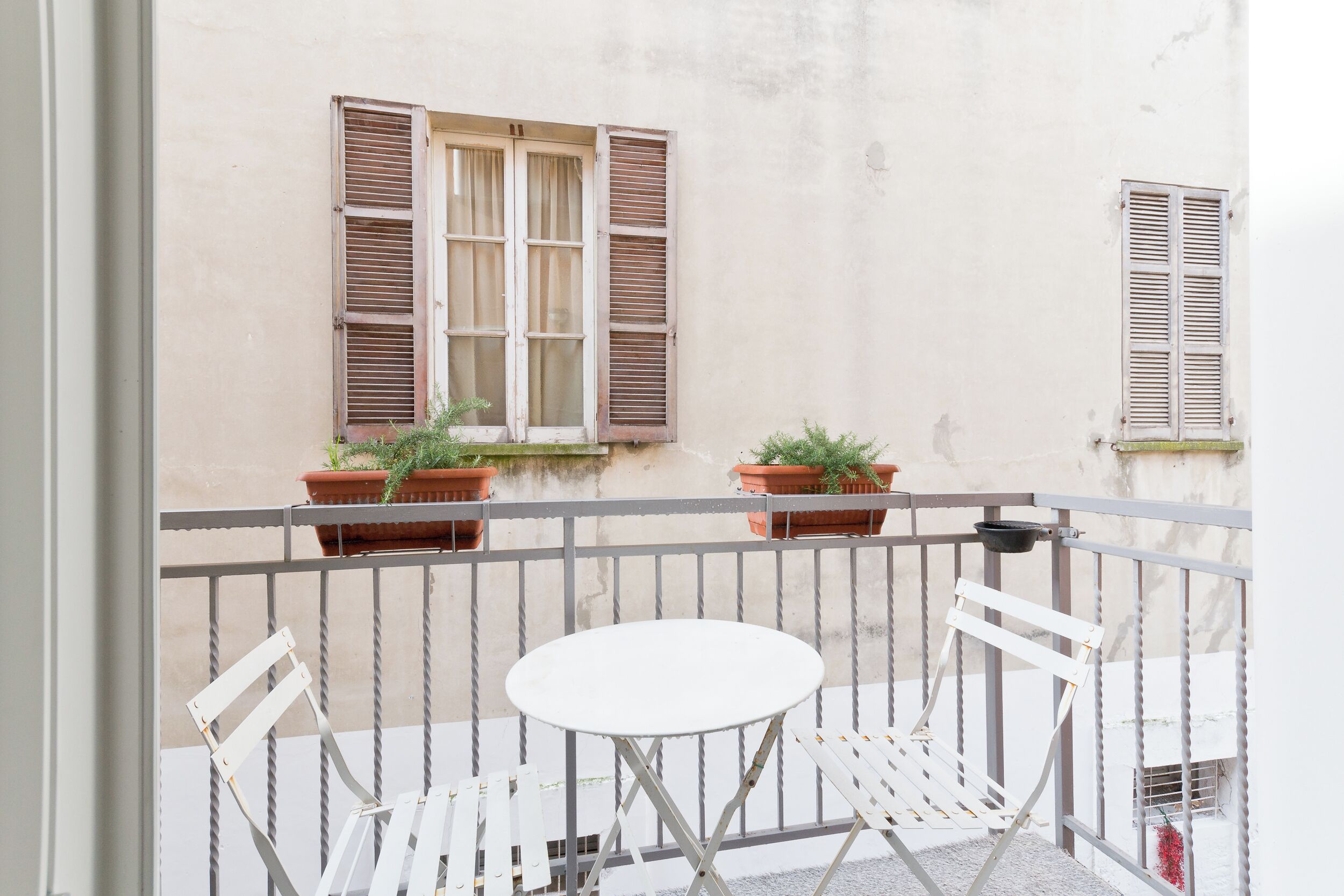 A balcony with a white table and two white chairs, potted plants, and a window with shutters.