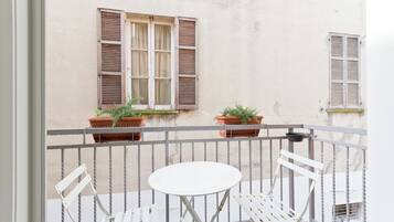 Ein Balkon mit weißem Tisch, zwei weißen Stühlen, Blumentöpfen und einem Fenster mit Rollläden.