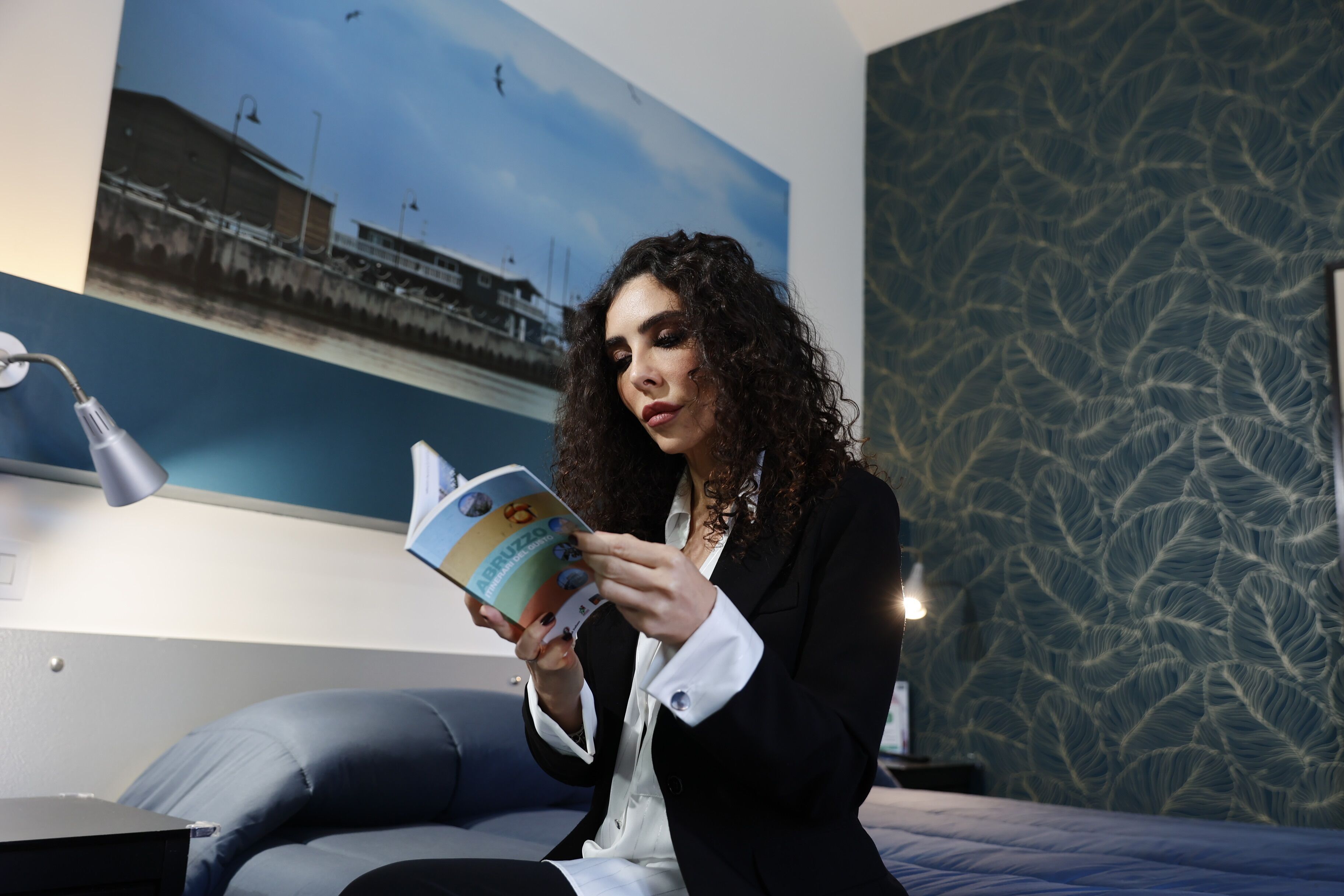 A person sitting on a bed in a room with a patterned wall, reading a book.
