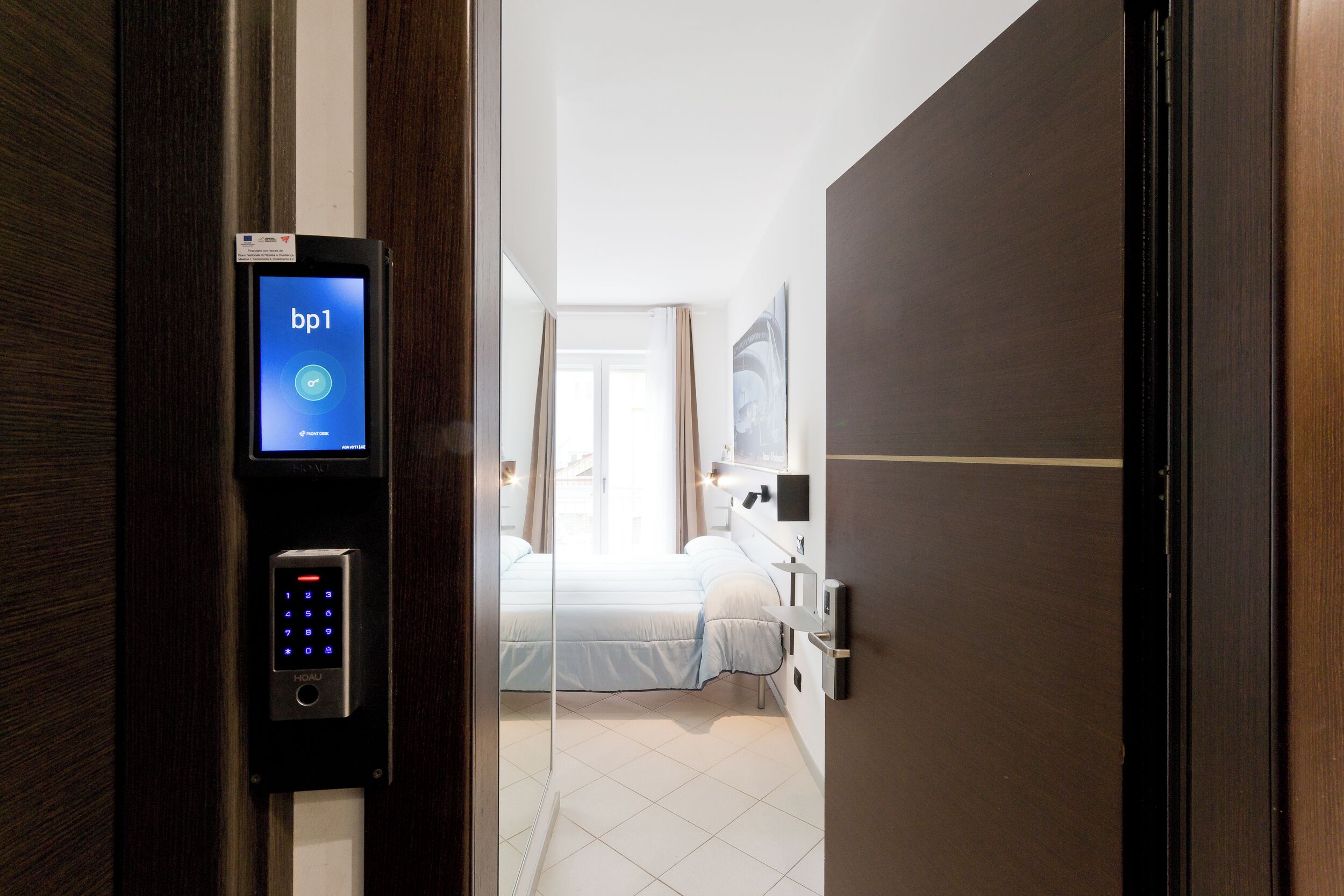 A hotel room with a digital keycard reader, a bed, a bedside table, and a window with curtains.