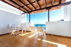 A balcony with a wooden ceiling, white chairs, a table, and a view of the ocean.