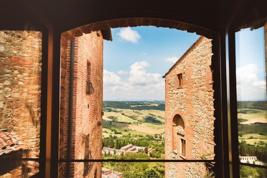 A view through a window of a stone building with a brick tower, overlooking a rural landscape with fields and a distant building.