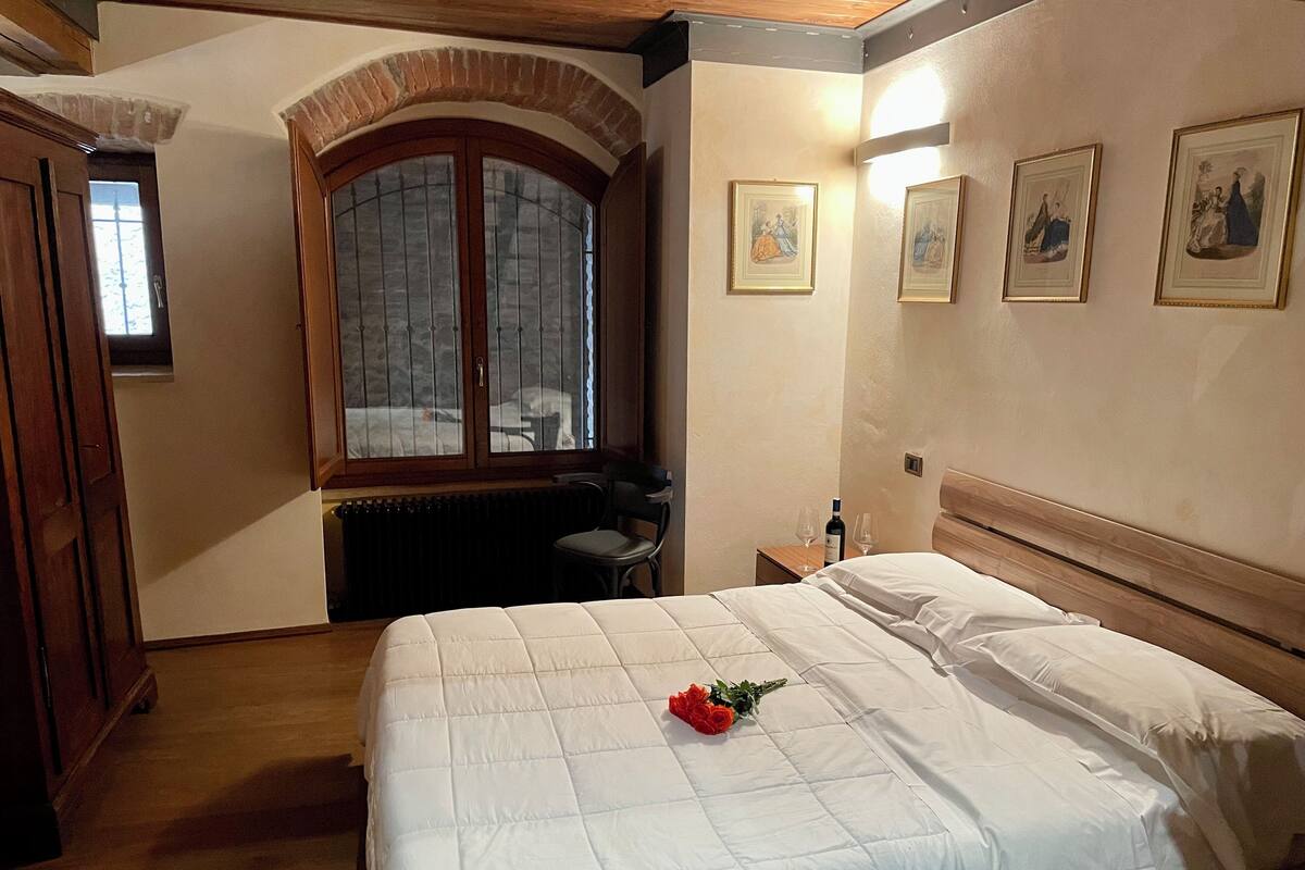 Habitación de hotel con una cama grande, una ventana arqueada de madera, un radiador, un escritorio y cuadros en la pared.
