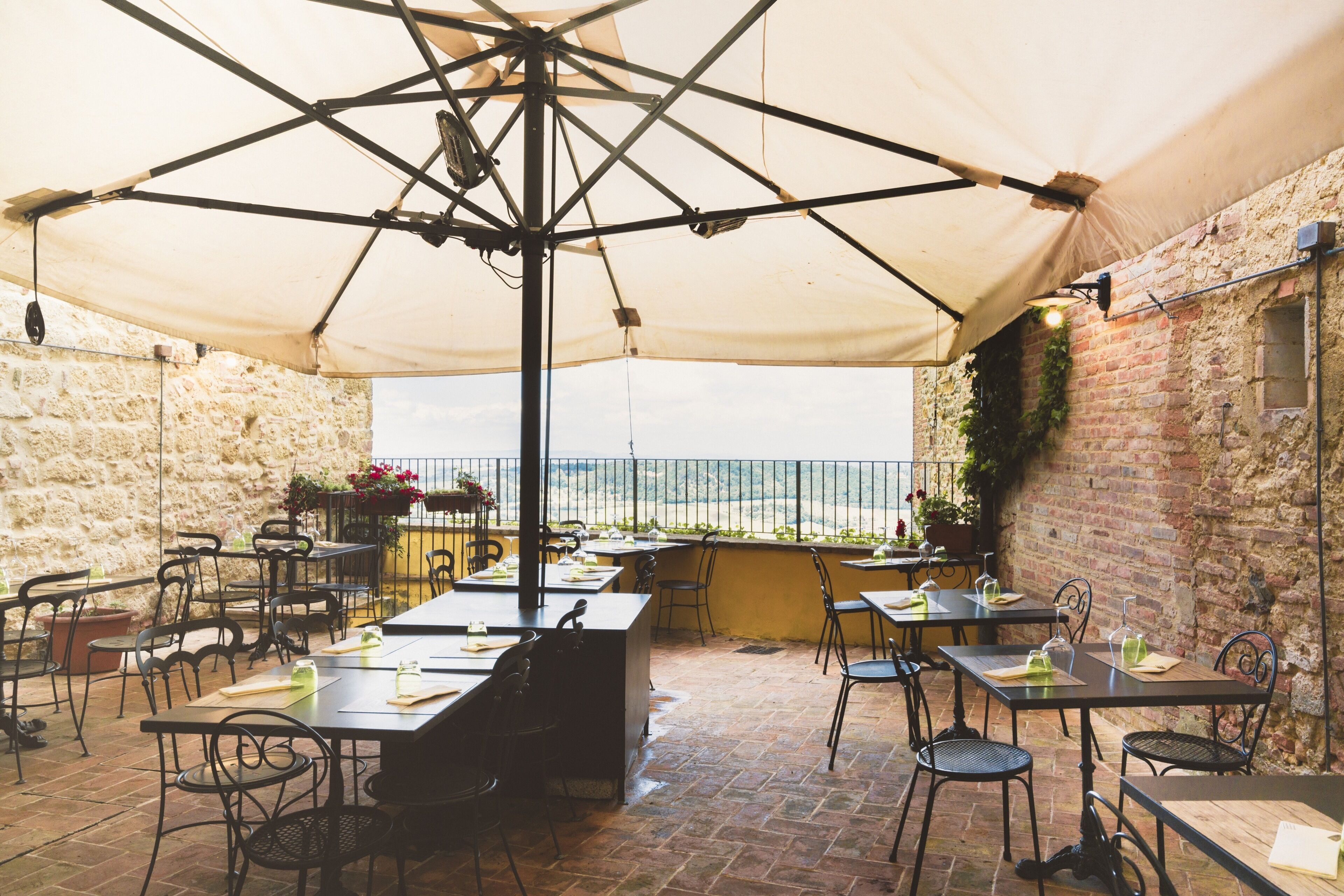 An outdoor patio with tables and chairs, a large umbrella, and a view of the sea.
