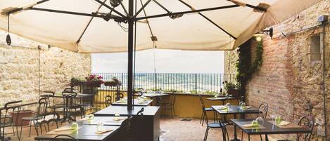 An outdoor patio with tables and chairs, a large umbrella, and a view of the sea.