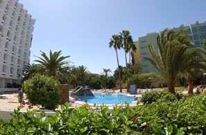 A pool area with palm trees, lounge chairs, and a building in the background. - HV Agaete Parque (San Bartolomé de Tirajana)