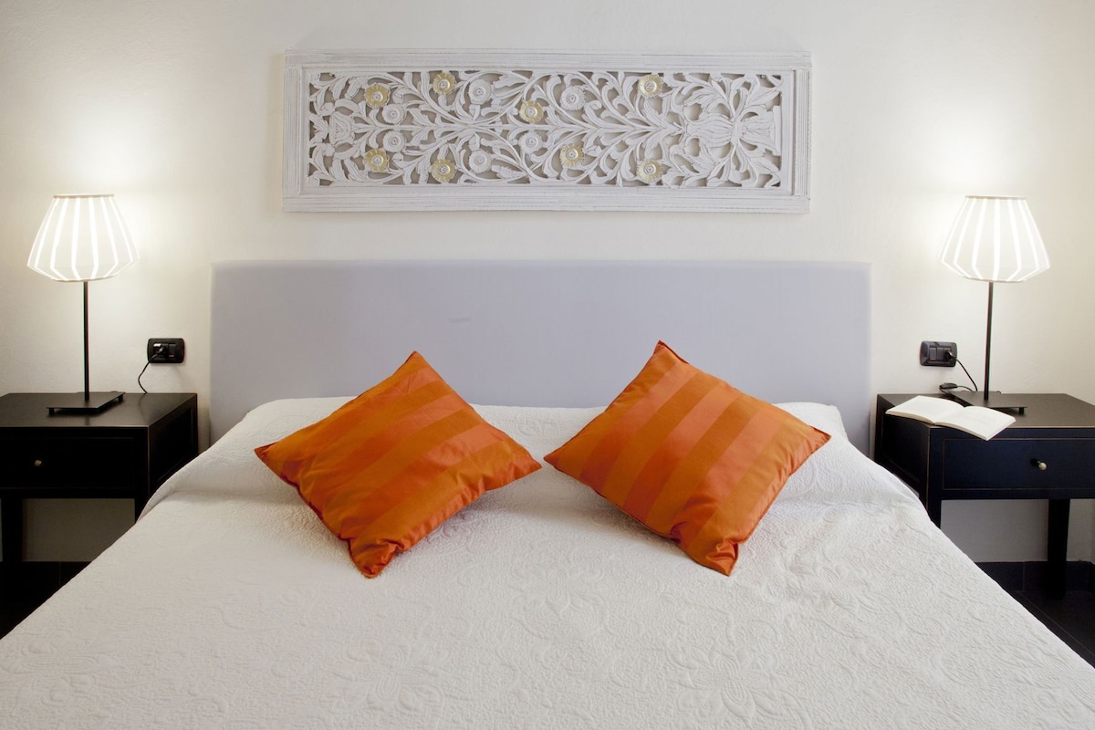 Una cama con ropa de cama blanca y dos almohadas naranjas, flanqueada por mesitas de noche con lámparas y un adorno decorativo en la pared superior.