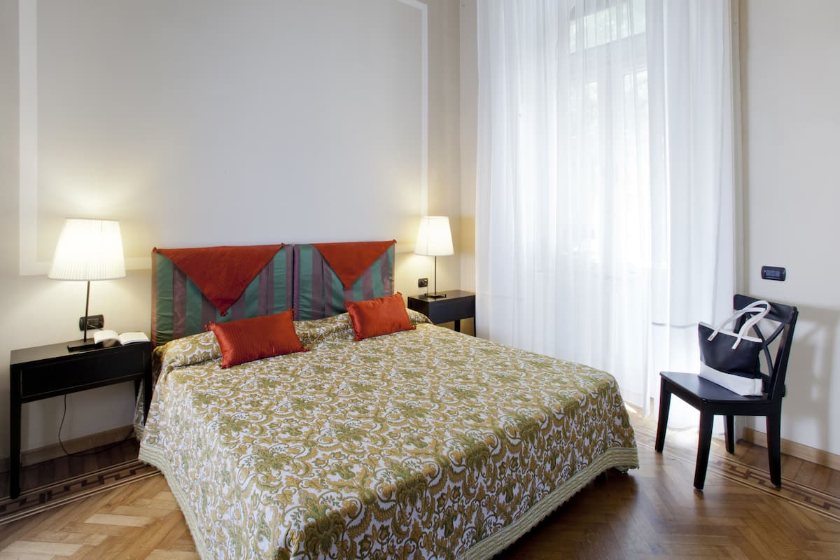 A hotel room with a large bed, two bedside tables with lamps, a chair, and a window with sheer curtains.