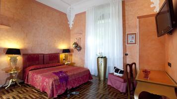 A hotel room with a large bed, two bedside lamps, a flat-screen TV, a desk with a chair, and a large potted plant.