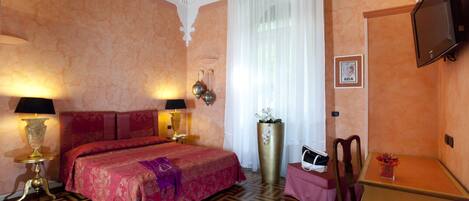 A hotel room with a large bed, two bedside lamps, a flat-screen TV, a desk with a chair, and a large potted plant.