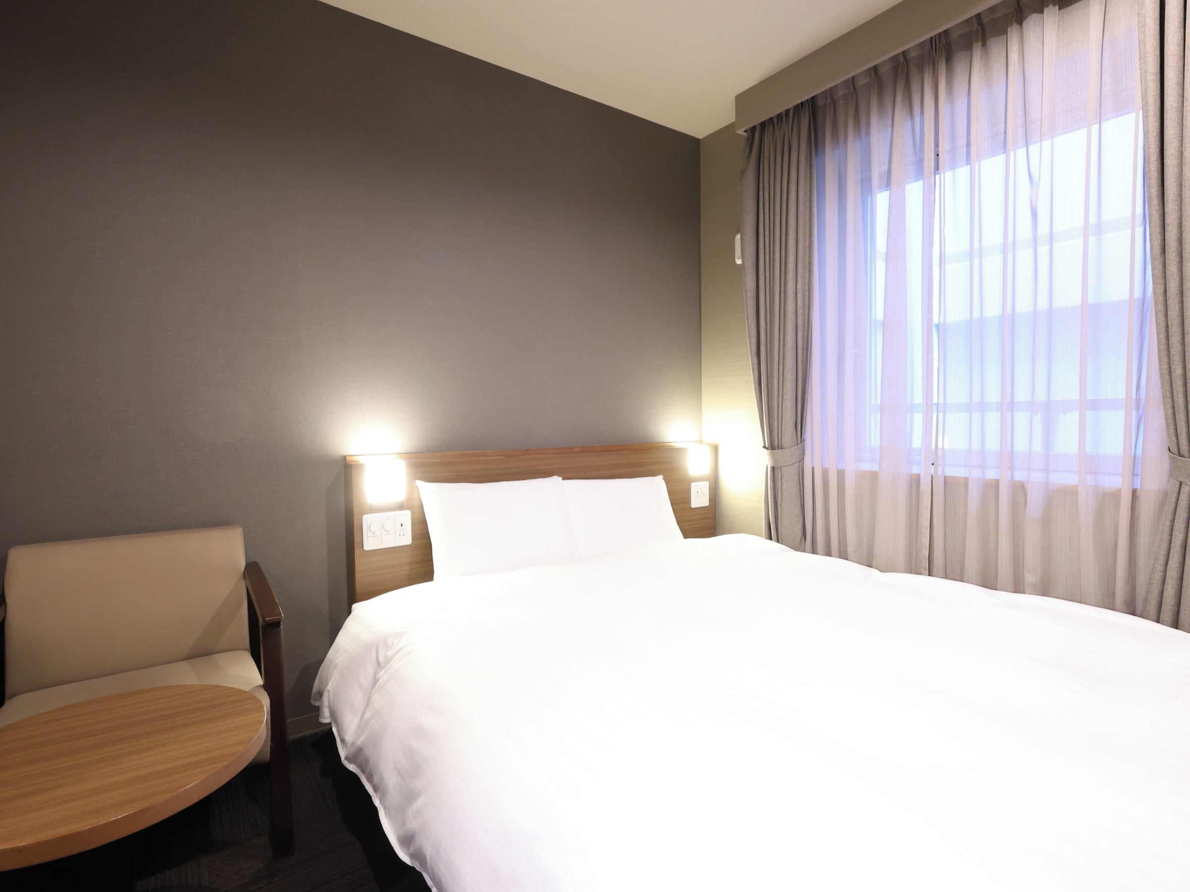 Quarto de hotel com cama, uma cadeira, uma mesinha e janela com cortinas.