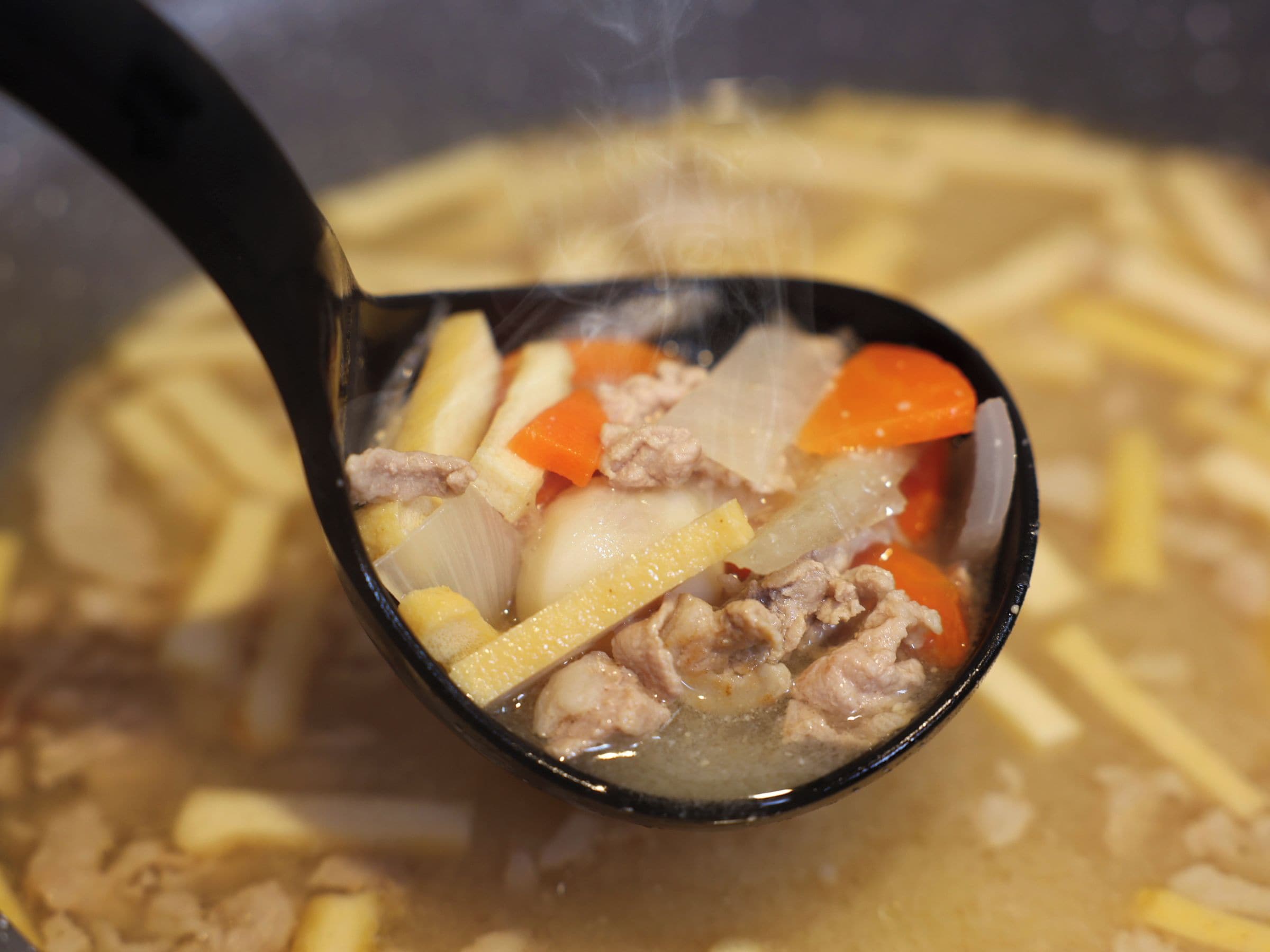 A close-up of a soup with visible ingredients like meat, carrots, and potatoes in a black bowl.