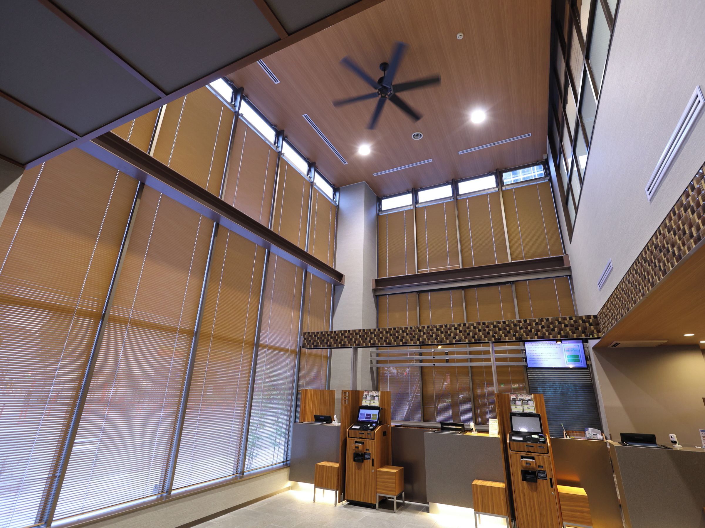 A spacious lobby with a high ceiling, large windows with blinds, a ceiling fan, and a reception desk with computers.