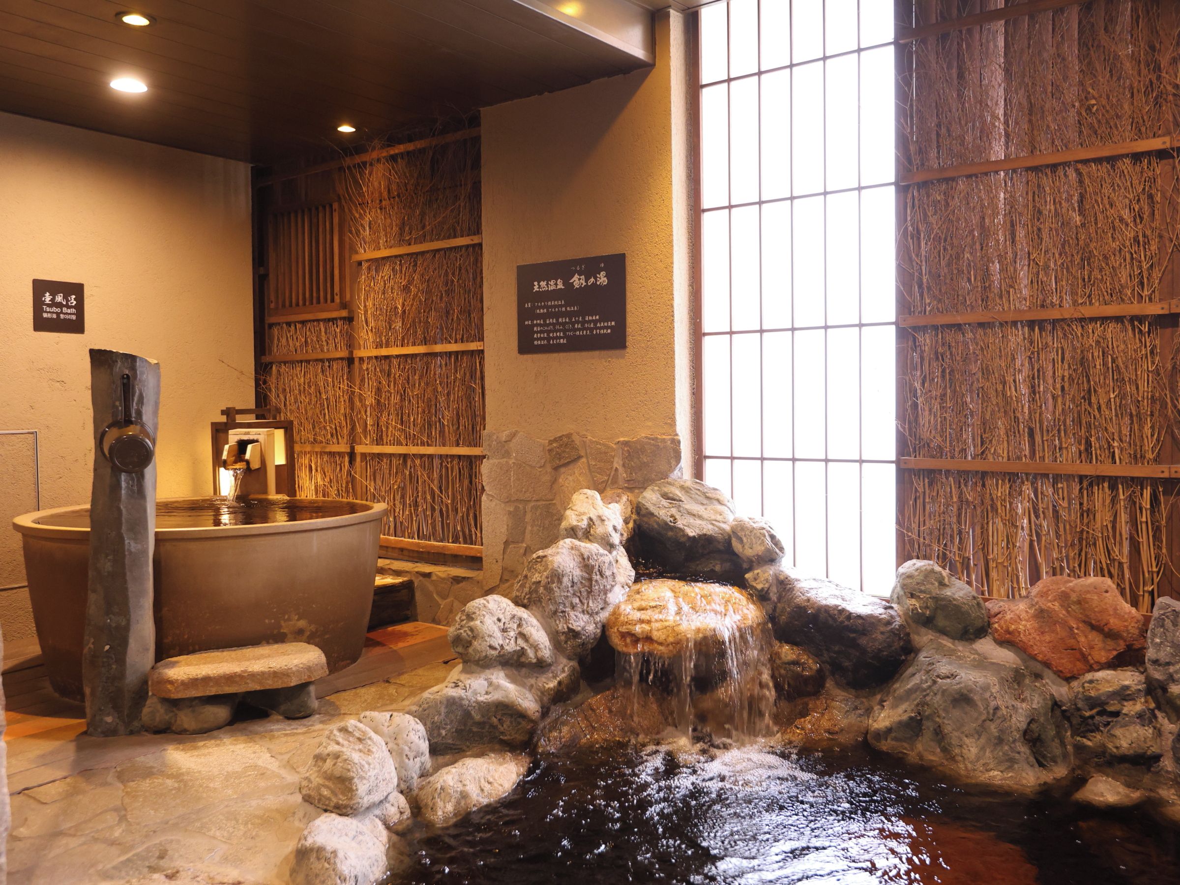 A traditional Japanese bath area with a stone basin, a wooden shelf with towels, and a rock waterfall feature.