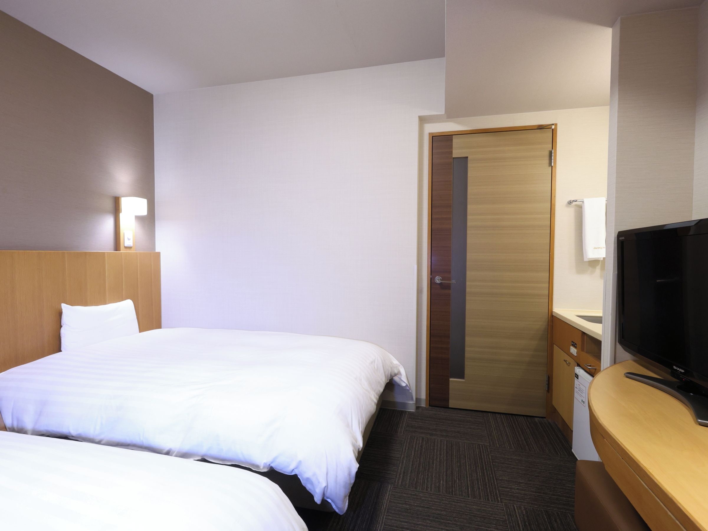 Habitación de hotel con dos camas, televisión, cabecero de madera, luz instalada en la pared y puerta que da a otra habitación.