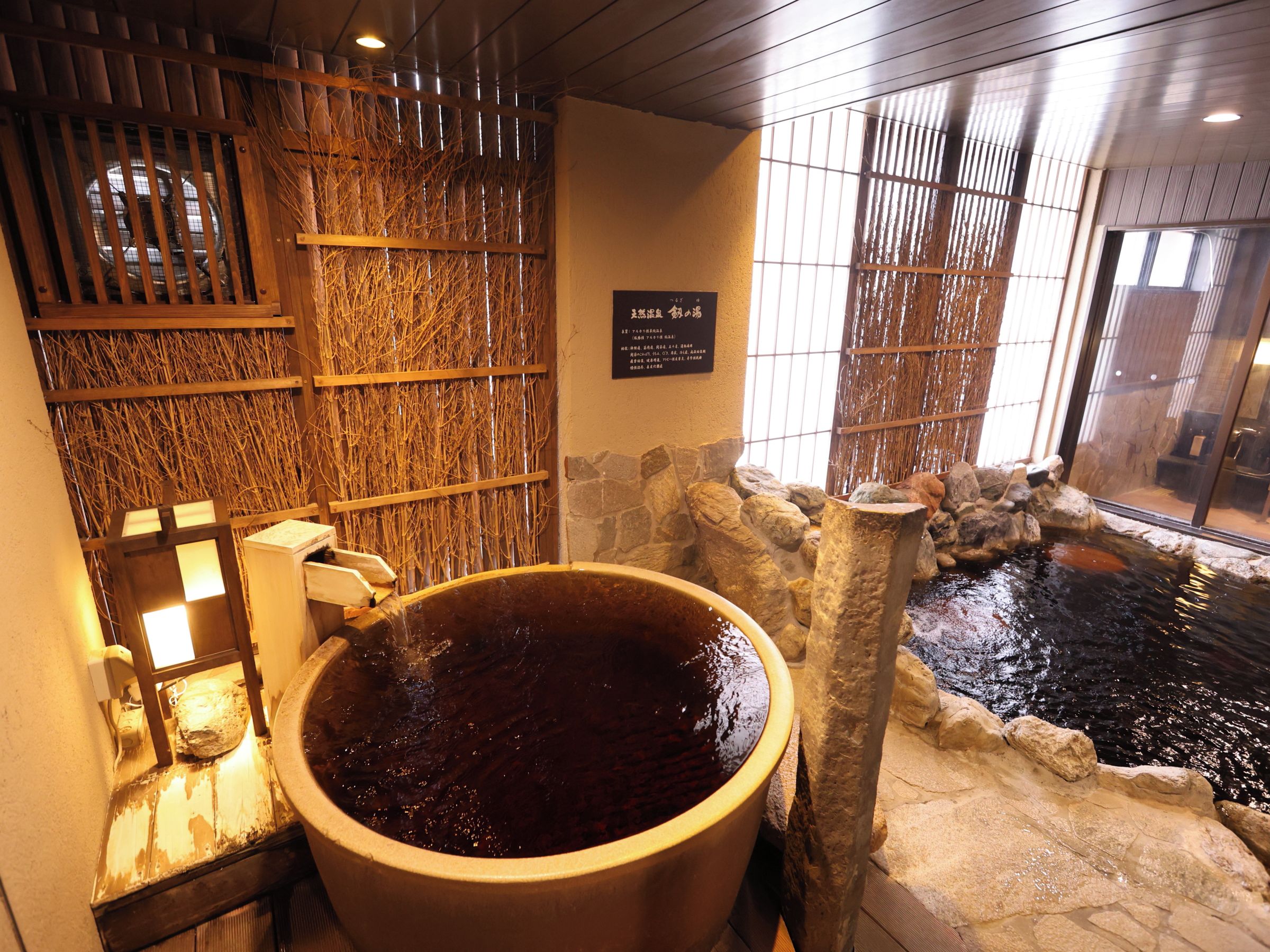 A hot spring bath area with a large stone basin, wooden partitions, and a small rocky pond.