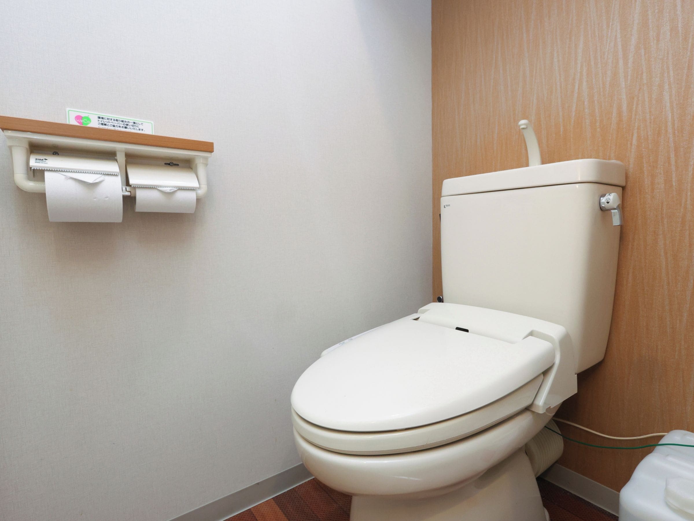 Des toilettes blanches, avec un réservoir et un abattant, un porte-rouleau de papier toilette contenant deux rouleaux, et un panneau en bois sur le mur.
