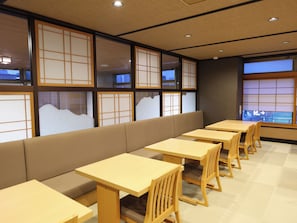 Un interno di un ristorante con tavoli e panche in legno, ampie finestre con paraventi di carta tradizionali e vista sull'esterno.