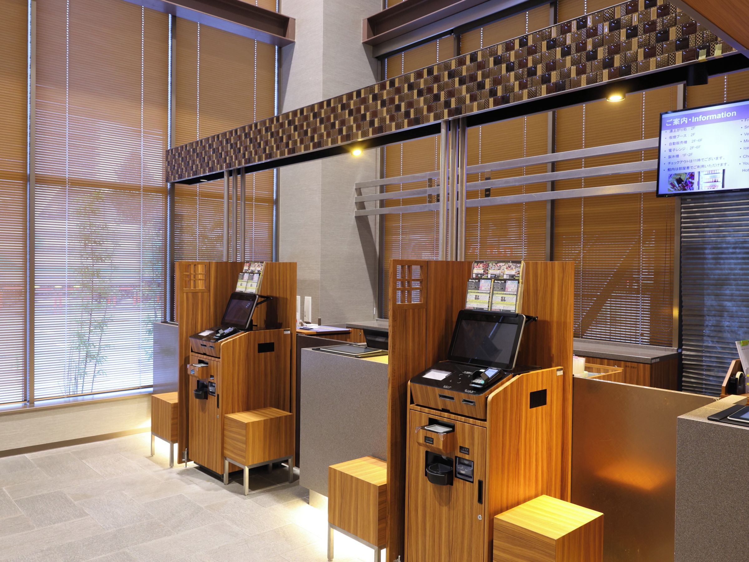 A modern hotel reception area with wooden reception desks, a digital information screen, and a view of the outside through large windows.