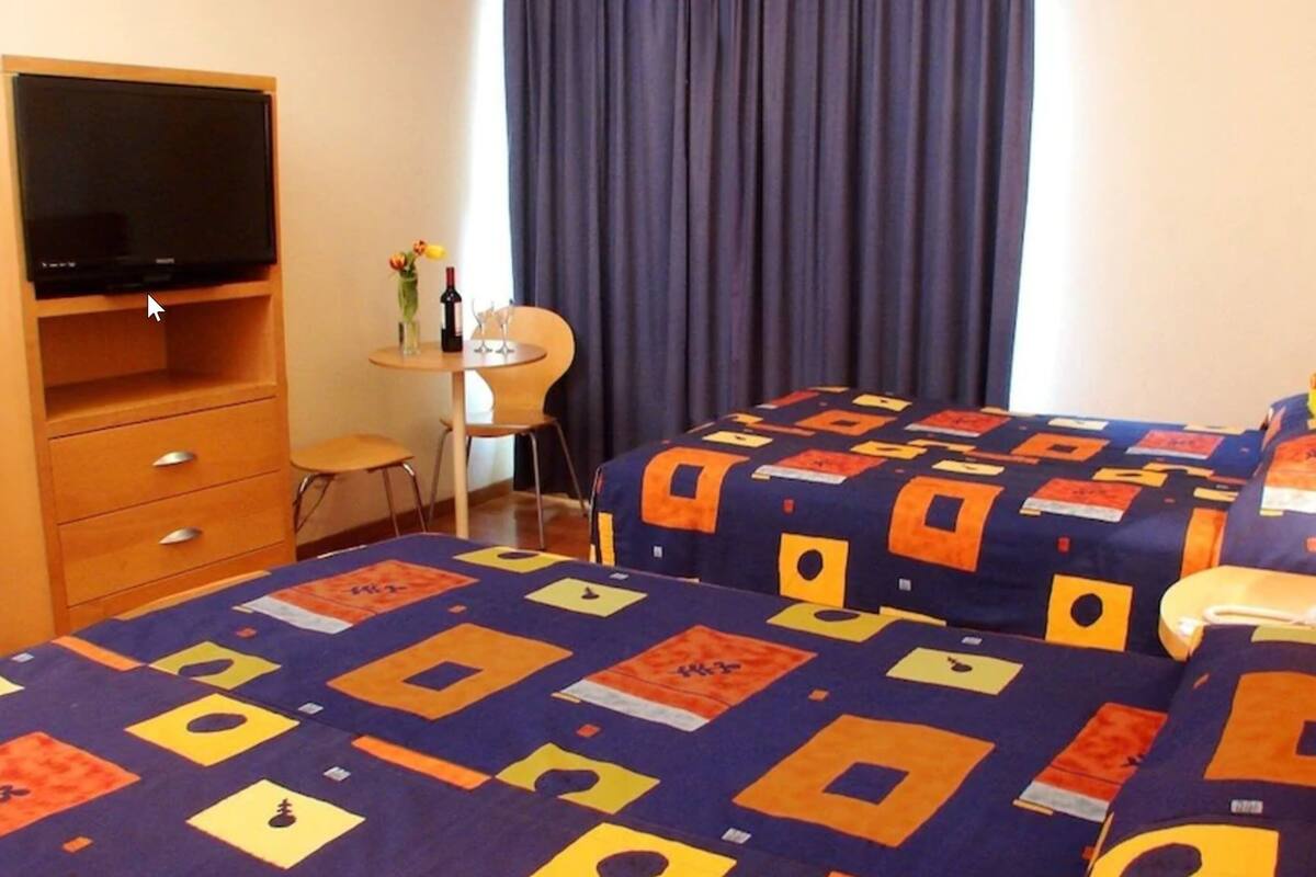 Quarto de hotel com duas camas, televisão, escrivaninha com cadeira e janela com cortinas.