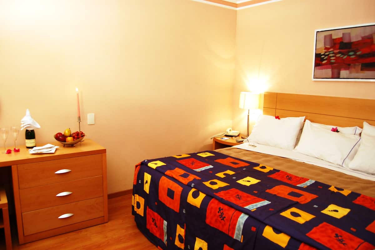 Quarto de hotel com cama, mesa de cabeceira, escrivaninha com gavetas, luminária, uma tigela de frutas e um quadro na parede.