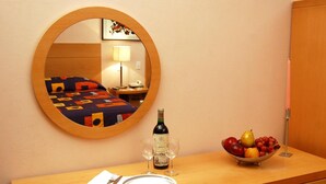 A hotel room with a round mirror, a bed with a colorful bedspread, a bedside table with a bottle of wine, two wine glasses, a bowl of fruit, and a neatly folded white napkin. - Hotel Del Principado (Mexico City)