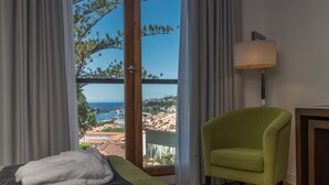 A hotel room with a bed, a green armchair, a lamp, and a view of the sea and buildings.