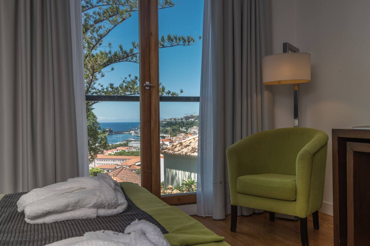 Habitación de hotel con cama, un sillón verde, una lámpara y vista al mar y edificios.