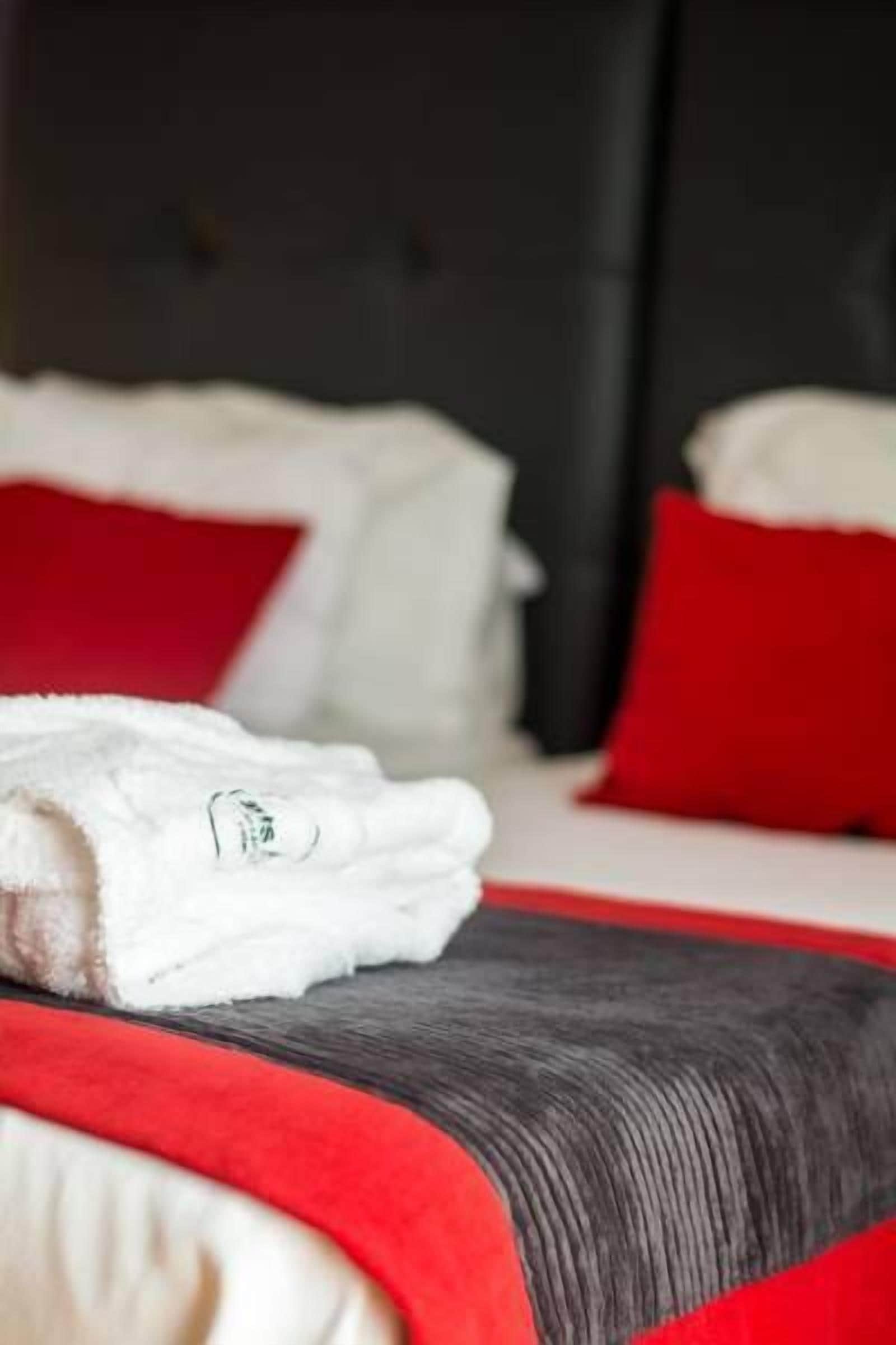 Un lit bien fait, avec du linge de lit blanc et des oreillers rouges, une tête de lit sombre et un coussin rouge décoratif posé sur le lit.